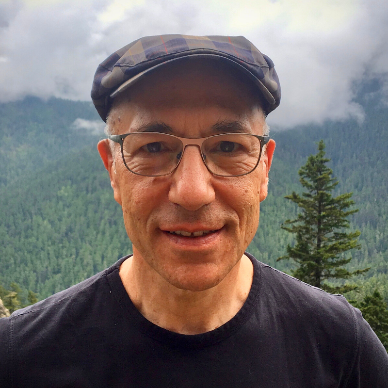 A fair-skinned man with light-rimmed glasses, black t-shirt, and flat cap stands in front of a mountain in the background.