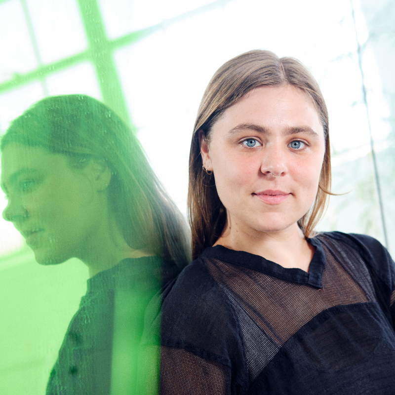 The artist, a white person, with blue eyes, and long blonde hair, reclines with their back against a mirror. They are wearing a black shirt, and a gold nose ring and earrings. The mirror behind them reflects their double in a green aura, also reflecting the sun's brilliance, a large plant growing, as well as factory windows, diffused in the background. The artist looks, almost smiling, directly into the camera.