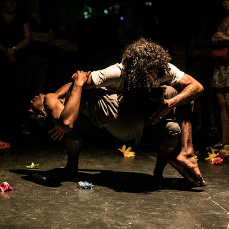 One dancer balances another on their lap on a stage with theatrical lighting