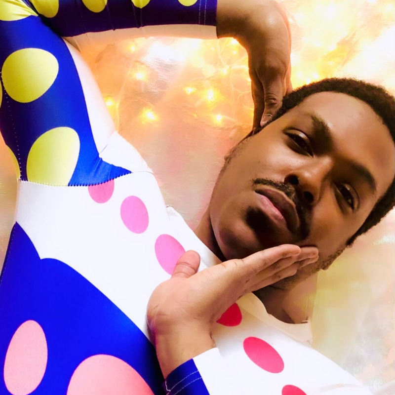 A dancemaker in a bodysuit with blue, white, and pink polka dots poses with his hands near his face.