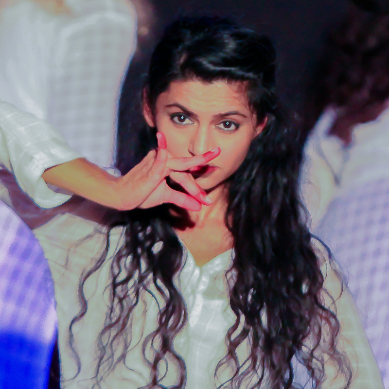 A dancemaker wearing soft blues and whites holds a hand gesture with flared fingers towards her mouth.