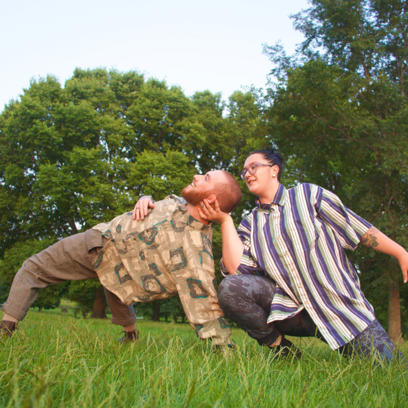 One white male dancer and one white non-binary dancer connect hand to head in an open field of green grass in the warm sunlight. He is wearing a brown, green, and yellow printed long sleeve button down, brown dance pants, and boots. They are wearing a white, blue and green vertically striped short sleeve button down shirt and printed blue dance pants, and black boots.