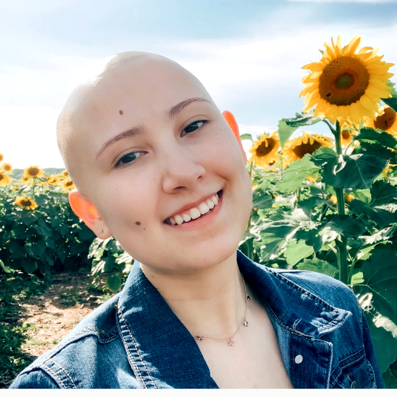 A headshot of an individua smiling in the sun with blue skies and a sunflower patch.