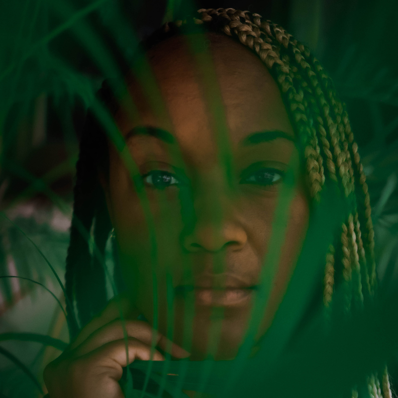 A Black woman peeking though the leaves of a bright green plant.