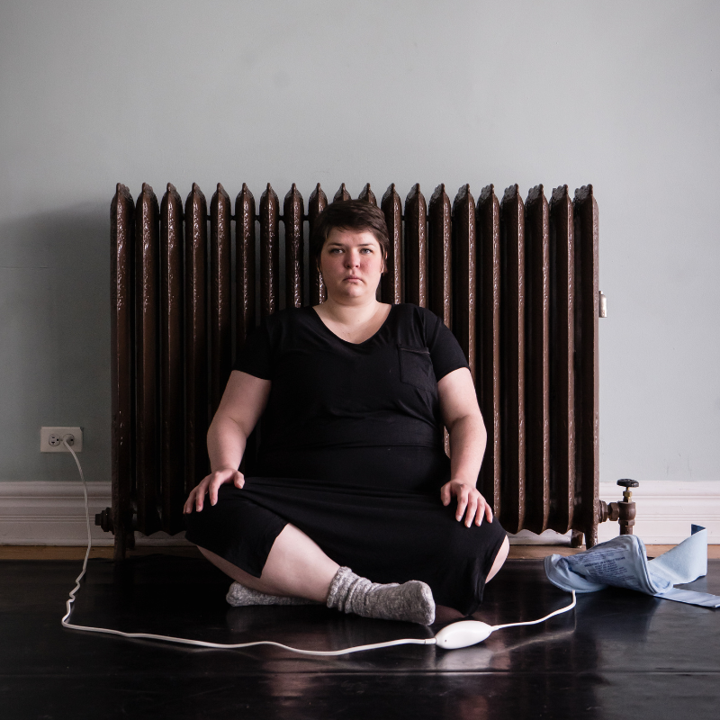 Maggie, a fat white woman with short brown hair, sits cross legged in front of a dark brown radiator against a light blue wall. She wears a long black nightgown. A heating pad lays on the floor beside Maggie, its cord stretched in a circle around her.