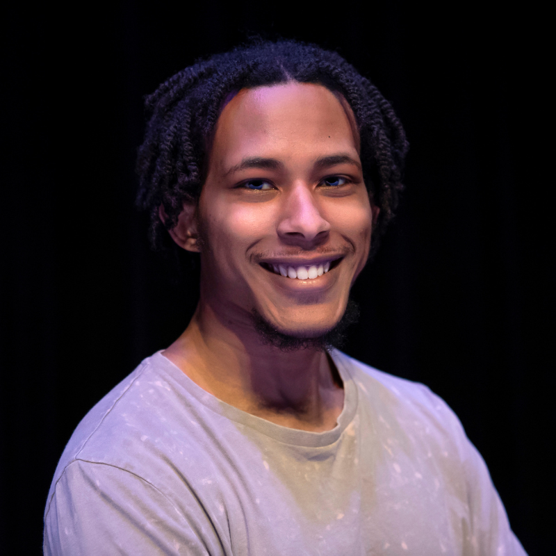 A headshot of an artist with brown skin smiling to the camera. He wears a pastel t-shirt and there is an effect of soft lighting. The background is black.